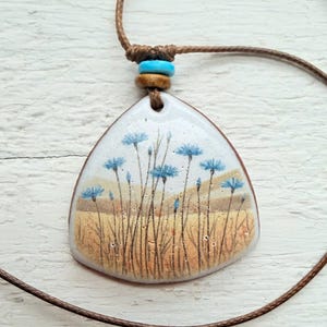 Puede incluir: Un collar con colgante con un colgante triangular de esmalte blanco. El colgante presenta un diseño floral pintado a mano con flores azules y hierba alta sobre un fondo beige y marrón. El collar tiene un cordón marrón con cuentas azules y marrones.