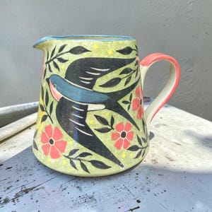 May include: A ceramic pitcher with a light green base featuring a black and blue swallow bird illustration, red flowers, and black leafy vines. The rim is trimmed in blue, and the handle is a solid coral pink color with a white interior.