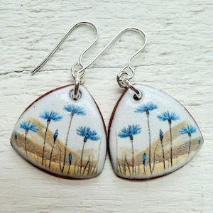 Puede incluir: Un par de pendientes hechos a mano con un diseño floral. Los pendientes tienen un fondo blanco con flores azules y detalles marrones. Los pendientes tienen forma triangular y tienen ganchos para las orejas plateados.