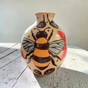 May include: A round ceramic vase with a cream-coloured base featuring a hand-painted bumblebee design in yellow, black, and light blue. The surface is decorated with black line-drawn leaves and red floral accents around the central bee motif.
