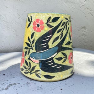 Peut inclure: Un gobelet en céramique tronconique au fond vert clair, orné d'une hirondelle bleue et noire peinte à la main en plein vol, entourée de branches feuillues vert foncé, de petits points jaunes et de fleurs corail à cinq pétales.