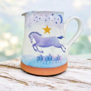 Puede incluir: Jarra de cerámica con un diseño caprichoso. La jarra presenta un caballo lavanda saltando sobre un cielo estrellado con una luna creciente y una estrella amarilla. La base es de color terracota y el asa es blanca.