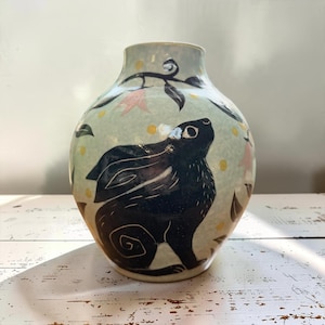 May include: A ceramic vase with a pale green background featuring a hand-painted black rabbit silhouette. The design includes small yellow dots, dark leaves, and soft pink floral accents. The vase has a rounded body and a narrow neck opening.