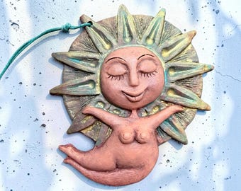May include: A terracotta-colored ceramic wall hanging featuring a sun face with closed eyes and a smiling mouth, surrounded by pointed rays with green and gold highlights. A figure with outstretched arms is attached to the lower sun rays.