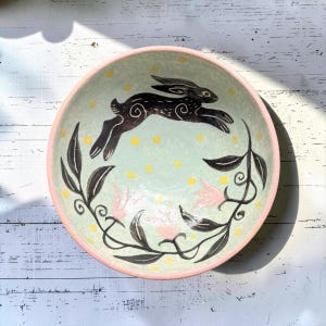 May include: A round ceramic bowl with a light green interior featuring a black leaping rabbit, black leafy vines, pink flowers, and small yellow dots. The bowl has a solid pink rim and a smooth, hand-painted finish throughout the interior surface.