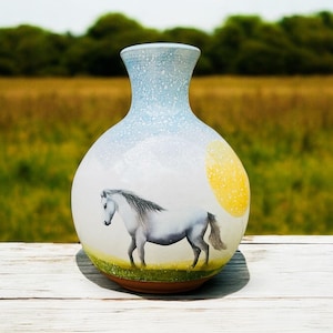 Könnte beinhalten: Keramikvase mit einer Illustration eines weißen Pferdes. Die Vase hat einen hellblauen, gesprenkelten oberen Teil, einen weißen Körper mit einer gelben Sonne und einen grün-braunen Sockel. Das Pferd ist grau und weiß.
