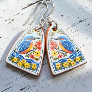 Könnte beinhalten: Ein Paar handgefertigte Ohrringe mit einem blauen und orangefarbenen Vogeldesign. Die Ohrringe haben einen weißen Hintergrund mit einem floralen Rand und silbernen Haken. Der Vogel sitzt auf einem Ast mit roten und gelben Blumen.