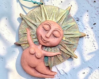 May include: A terracotta-colored ceramic wall hanging featuring a central sun face with closed eyes and a smiling expression, surrounded by radiating pointed rays. A figure with a rounded body is positioned in front of the sun, suspended by a teal cord.