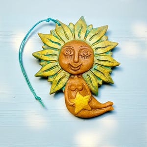 以下が含まれることがあります： 笑顔の太陽人魚の陶器製手作り置物。黄色い星を持っています。太陽の花びらは黄色と緑色で、吊り下げ用のターコイズ色の紐が付いています。置物の高さは約15cmです。