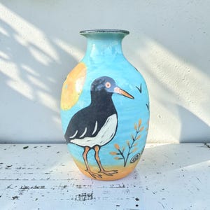 May include: A ceramic vase with a light blue background featuring a painted black and white bird with an orange beak and legs. The design includes a large yellow sun, small black bird silhouettes, and orange plant sprigs near the base.