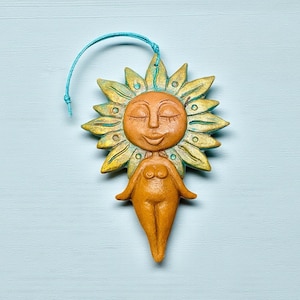 以下が含まれることがあります： 笑顔で目を閉じた陶器製の太陽の女神のオーナメント。太陽の光線は黄色と緑で、体はオレンジ色です。吊り下げ用の青い紐が付いています。