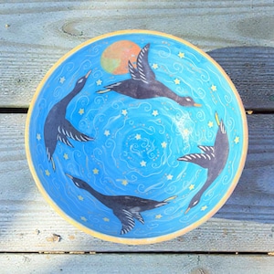 May include: A hand-painted ceramic bowl with a blue interior featuring four black birds in flight, a yellow sun, and white stars. The bowl has a yellow rim and is set against a wooden surface.