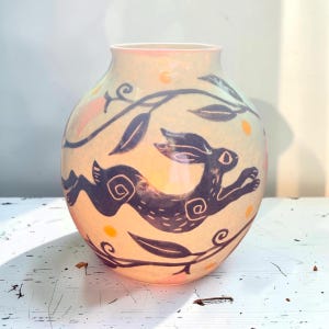 May include: A round ceramic vase with a pale peach glaze, featuring a dark purple hand-painted illustration of a leaping hare surrounded by stylized leafy vines and small yellow dots. The vase has a smooth finish and a wide opening.