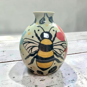 以下が含まれることがあります： クリーム色のベースに、黄色と黒で手描きされた大きなマルハナバチが描かれた丸い陶器の花瓶。デザインには、小さな黒い葉の輪郭、散らばった黄色い点、そして表面全体に柔らかな赤と青のアクセントが含まれています。