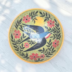 May include: A round ceramic bowl with a yellow rim, featuring a central blue and black swallow bird illustration. The background is a mottled light green with a yellow sun, surrounded by a decorative pattern of black leafy vines and red flowers.