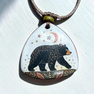 May include: A triangular pendant on a brown cord featuring a black bear walking across a forest floor with oak leaves and berries. The background is white with a crescent moon and small stars above the bear. The pendant has a green bead accent.