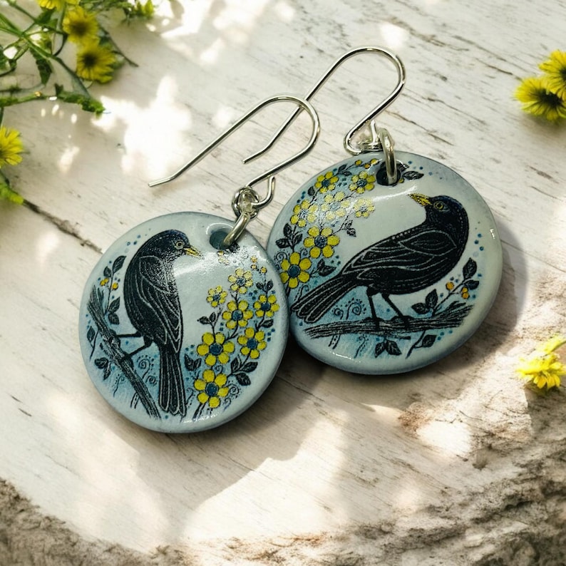 Pu&ograve; includere: Un paio di orecchini pendenti in argento con ciondoli rotondi in ceramica blu e bianca. Ogni ciondolo presenta un uccello nero appollaiato su un ramo con fiori gialli.
