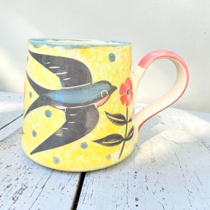 May include: A ceramic mug with a bright yellow background featuring a dark blue swallow in flight, small blue polka dots, and a red flower with green leaves. The mug has a curved handle with a pinkish-red accent on the outer edge and rim.
