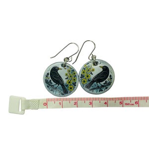 Pu&ograve; includere: Un paio di orecchini pendenti in argento con ciondoli rotondi in ceramica blu e bianca con uccelli neri e fiori gialli. Gli orecchini sono lunghi 5 cm.