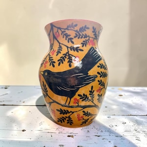 May include: A ceramic vase with a warm yellow-orange base color, featuring a hand-painted black bird perched on a branch. The design includes dark brown leafy branches and small clusters of red berries scattered across the surface of the vase.