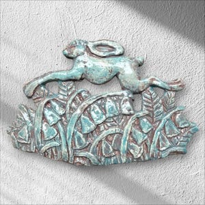 May include: A decorative ceramic wall plaque featuring a relief sculpture of a leaping hare positioned above a cluster of bell-shaped flowers and foliage. The piece has a glossy, mottled teal and blue glaze with visible brown clay undertones.