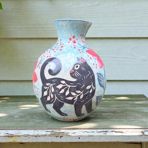 Puede incluir: Un jarrón de cerámica con un fondo azul claro y un diseño de gato negro. El gato está decorado con patrones de hojas blancas. Acentos florales rojos y un borde azul completan el diseño. El jarrón está sobre una superficie de madera.