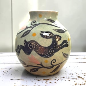 Könnte beinhalten: Eine runde Keramikvase mit hellgrüner Glasur, die einen handgemalten, dunkelbraunen Hasen in einer Sprungbewegung zeigt. Das Design umfasst schwarze Blatt-Rankenmuster, kleine gelbe Punkte und zarte rosa Blumenakzente.