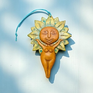 以下が含まれることがあります： 太陽の女神のセラミック壁掛け。笑顔の顔と両手を広げた姿で、ひまわりのような光輪があります。色は茶色、金色、緑色のアースカラーで、吊り下げ用の青い紐が付いています。ユニークなアート作品です。