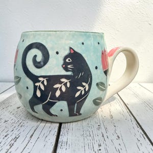 Op de afbeelding: Een lichtblauwe keramische mok met een illustratie van een zwarte kat met witte bladpatronen op het lichaam. De kat heeft een gekrulde staart en is omringd door kleine zwarte stippen en roze bloemen. De mok heeft een gebogen handvat en een ronde vorm.