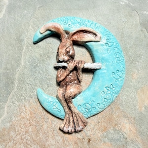 May include: A ceramic sculpture of a brown rabbit playing a flute while seated on a light blue crescent moon. The moon has a floral pattern. The sculpture is on a grey stone surface.
