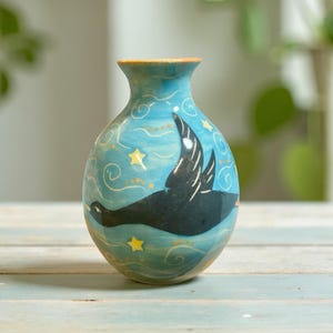 Peut inclure: Un vase en céramique avec un fond bleu clair, représentant un oiseau noir en vol, des étoiles jaunes et des volutes blanches. Le vase a un col étroit et un corps arrondi, avec un bord orange.