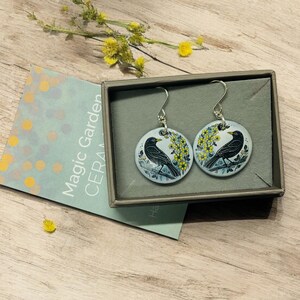 Pu&ograve; includere: Un paio di orecchini d'argento con un ciondolo in ceramica blu e bianca che presenta un merlo appollaiato su un ramo con fiori gialli. Gli orecchini sono in una piccola scatola regalo con le parole "Magic Garden Ceramics" sul lato.