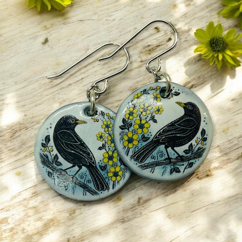 Pu&ograve; includere: Un paio di orecchini pendenti in argento con un ciondolo rotondo in ceramica che presenta un merlo appollaiato su un ramo con fiori gialli. Lo sfondo del ciondolo &egrave; di colore azzurro chiaro.