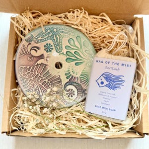 May include: A ceramic soap dish featuring a seahorse and sea life design in green, purple, and beige, alongside a bar of goat milk soap. The soap is labelled "HAG OF THE MIST Lost Lands". The soap weighs 85g.