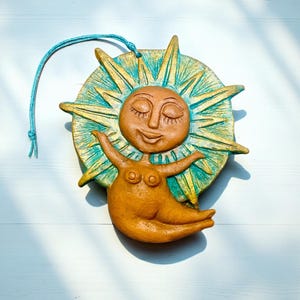 以下が含まれることがあります： 笑顔と両手を広げた陶器製の太陽の置物。太陽は茶色の顔と体をしており、周りにはターコイズとゴールドのサンバーストのデザインが施されています。吊り下げ用の青い紐が付いています。