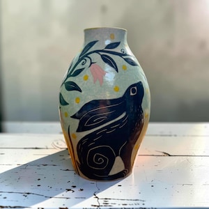 May include: A ceramic vase with a light blue background featuring a large black rabbit illustration. The design includes dark green leafy branches, a single pink bell-shaped flower, and scattered yellow dots across the surface.
