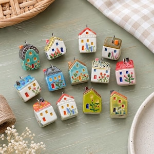May include: A collection of thirteen small, three-dimensional ceramic house charms, each featuring a unique hand-painted design with colourful roofs, windows, and floral details. Each charm includes a metal loop at the top for hanging.