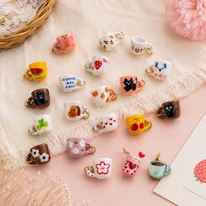 May include: A collection of miniature ceramic mug charms featuring various colorful designs including animals, fruits, flowers, and text. Each small mug includes a gold-tone metal jump ring attached to the handle for use as a jewelry pendant.