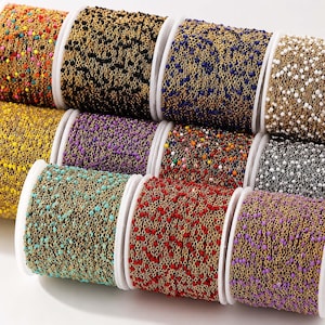 Puede incluir: Una colección de once bobinas de cadena de metal dorado, cada una con pequeñas cuentas de esmalte espaciadas uniformemente en varios colores sólidos, incluyendo rojo, azul, negro, blanco, amarillo, morado, turquesa y una variedad multicolor.