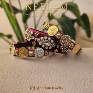 Peut inclure: Deux bracelets rigides en bordeaux profond enveloppés de fil de soie, ornés de breloques dorées en forme de pièces, de cristaux facettés transparents et de petites perles blanches. Le texte NEELAM JEWELRY, @neelam.jewelry et neelamjewelryfr.etsy.com est visible.