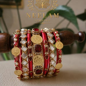 Peut inclure: Un ensemble de bracelets joncs en fil de soie rouge, ornés d'accents en métal doré, de pierres transparentes en forme de losange et de pièces de monnaie dorées. Le bracelet central présente des pierres rouges rectangulaires et le logo NEELAM JEWELRY.