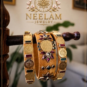 Può includere: Tre bracciali rigidi dorati con superficie in tessuto testurizzato, decorati con ciondoli metallici a forma di moneta e pietre preziose rettangolari e floreali nei toni del rosso scuro, verde e trasparente. Il marchio NEELAM JEWELRY è visibile sopra.