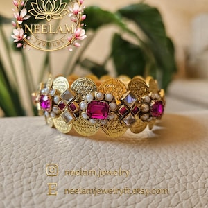 Peut inclure: Un bracelet jonc doré composé d'une série de disques circulaires semblables à des pièces de monnaie, agrémenté de pierres rectangulaires rose fuchsia, de pierres transparentes en losange et de petites perles blanches. Texte : NEELAM JEWELRY, neelam.jewelry, neelamjewelryfr.etsy.com.