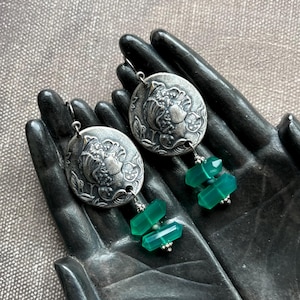 May include: Silver-toned dangle earrings. Each earring features a round, embossed pendant with a floral design and a cluster of green, faceted gemstones. The earrings are displayed on a black hand-shaped sculpture against a textured background.