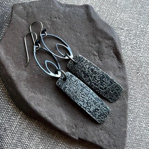 May include: A pair of silver earrings with a textured rectangular pendant and a delicate openwork design. The earrings are displayed on a dark gray stone surface.