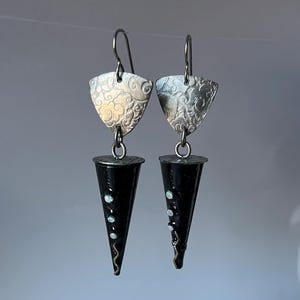May include: A pair of handcrafted earrings. Each earring features a silver, shield-shaped top with an embossed floral design, connected to a black, cone-shaped bottom adorned with white dots and a wavy silver line. The earrings hang from silver hooks.