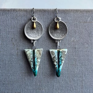 May include: A pair of handmade earrings. Each earring features a silver coin with a gold-colored bead, connected to a triangular pendant with blue, teal, and gray patterns. The earrings hang from silver hooks.