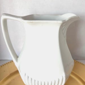May include: A white ceramic pitcher featuring a curved handle, a pouring spout, and a base decorated with vertical fluted ridges. The body of the pitcher has a smooth, solid white finish with subtle contours and a square-shaped bottom edge.