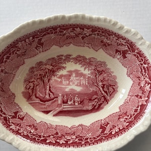 May include: An oval ceramic bowl with a cream-colored base and a deep red transferware pattern. The center features a detailed landscape with a castle and figures, while the rim is decorated with a dense border of intricate grape leaves and vines.