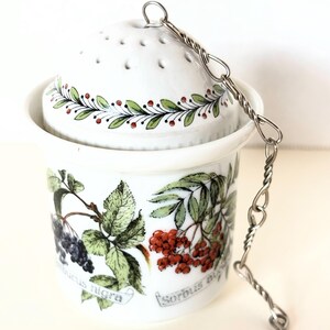 May include: A white ceramic tea infuser with a perforated dome lid and a metal chain. The body features botanical illustrations of berries and leaves with the text Sambucus nigra and Sorbus aucuparia printed in black script near the base.